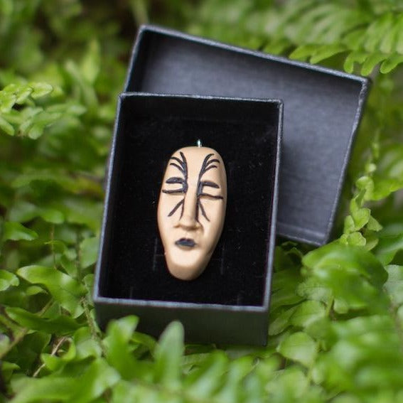 Light brown face form pendant with black lines imitating maori style tattoos. Pendant placed in black gift box surrounded by green ferns