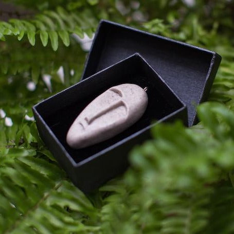 Beige colour tribal mask pendant in black gift box and green fern background 
