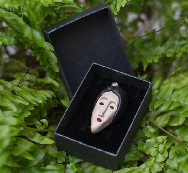 Face pendant that made from polymer clay looks like Modigliani's woman's portrait is in black gift box surrounded by green ferns