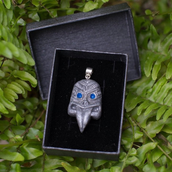 Grey maori style tribal mask charm (with sticking out tongue and blue eyes) in black gift box and placed in green fern background  
