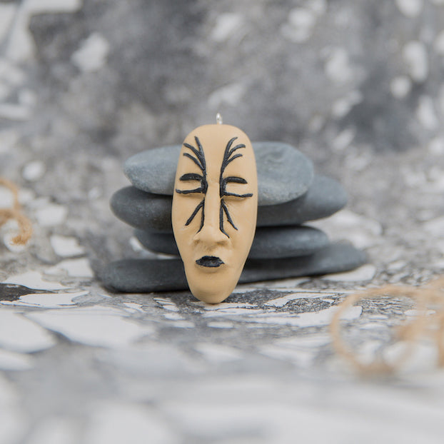 Hand-sculpted face shape pendant with black lips and closed eyes and tattoo lines around nose. Pendant is placed in grey neutral background
