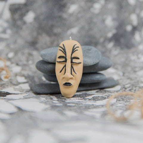 Hand-sculpted face shape pendant with black lips and closed eyes and tattoo lines around nose. Pendant is placed in grey neutral background
