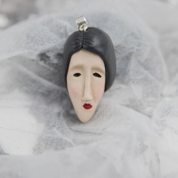 Hand-sculpted from polymer clay woman's face pendant with black hair and red lips looks like Modigliani style painting lying on white background