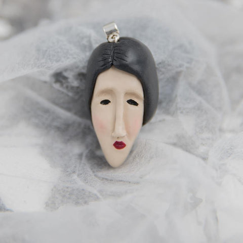 Hand-sculpted from polymer clay woman's face pendant with black hair and red lips looks like Modigliani style painting lying on white background